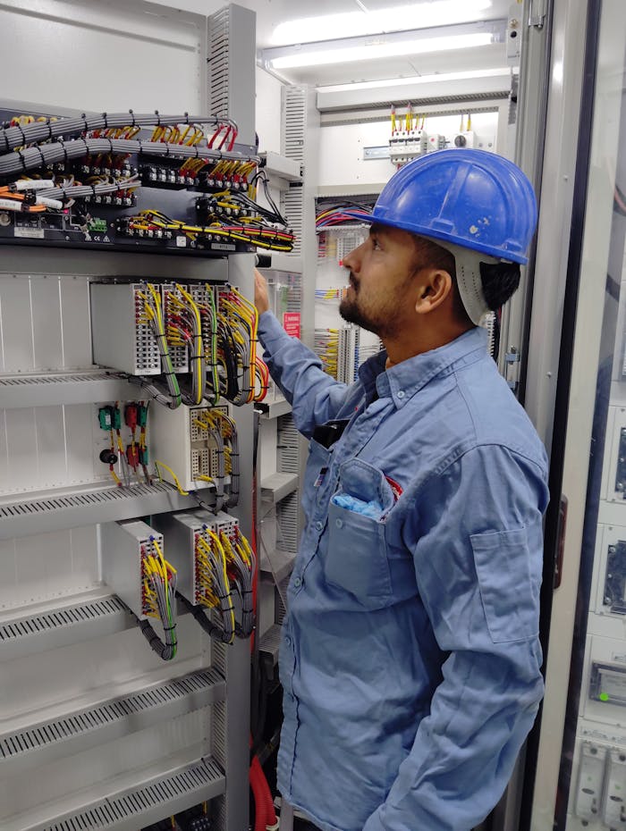 Electrician in blue helmet checking control panel wiring for safety compliance.